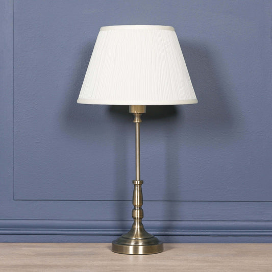 Traditional satin brass candlestick table lamp with cream pleated shade on a blue wall.