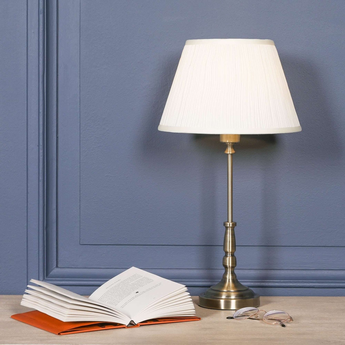Traditional satin brass candlestick metal table lamp with cream shade beside open book and glasses.