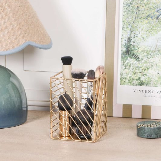 Gold Painted Square Metal Storage Basket holding makeup brushes on a wooden table beside a lamp and artwork.