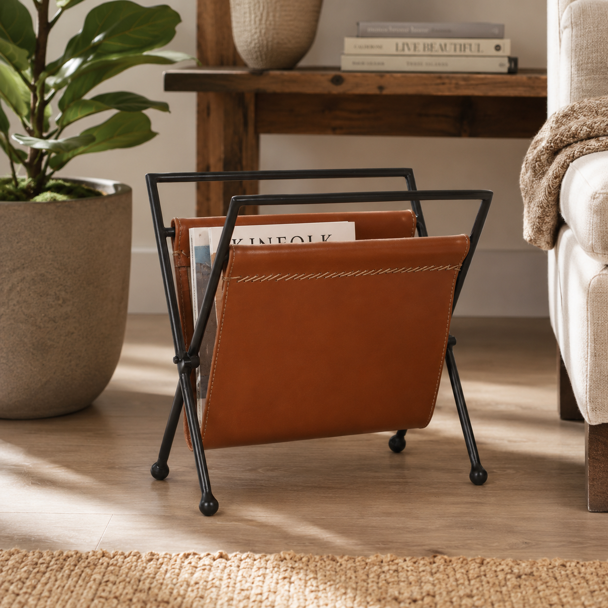 Brown leather magazine holder with black metal frame on a wooden floor, next to a plant and a sofa.