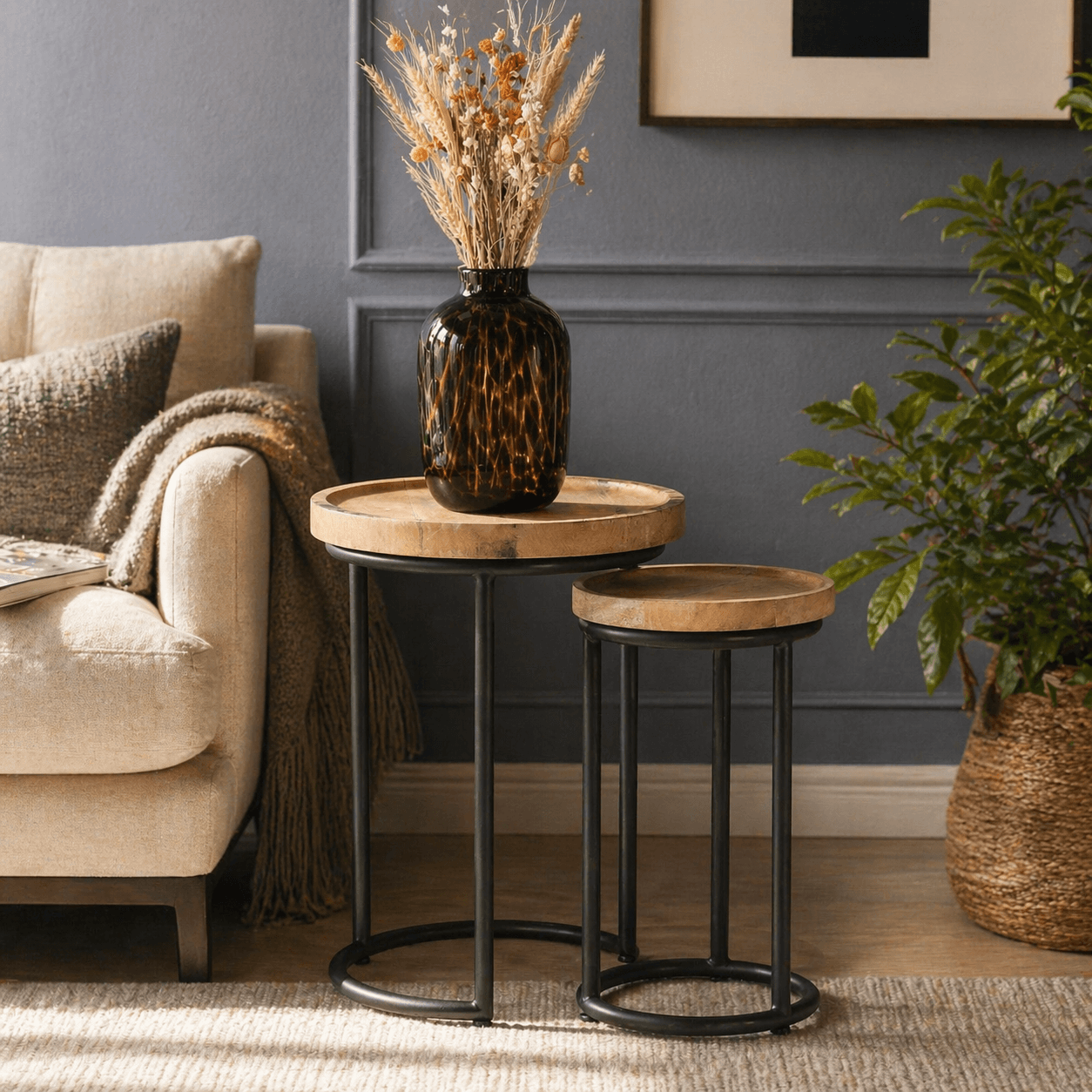 Modern nested side tables with round wooden tops and black metal frames styled beside a beige sofa in a cozy living room, featuring a dark glass vase with dried flowers, soft textiles, and a leafy plant against a blue paneled wall.