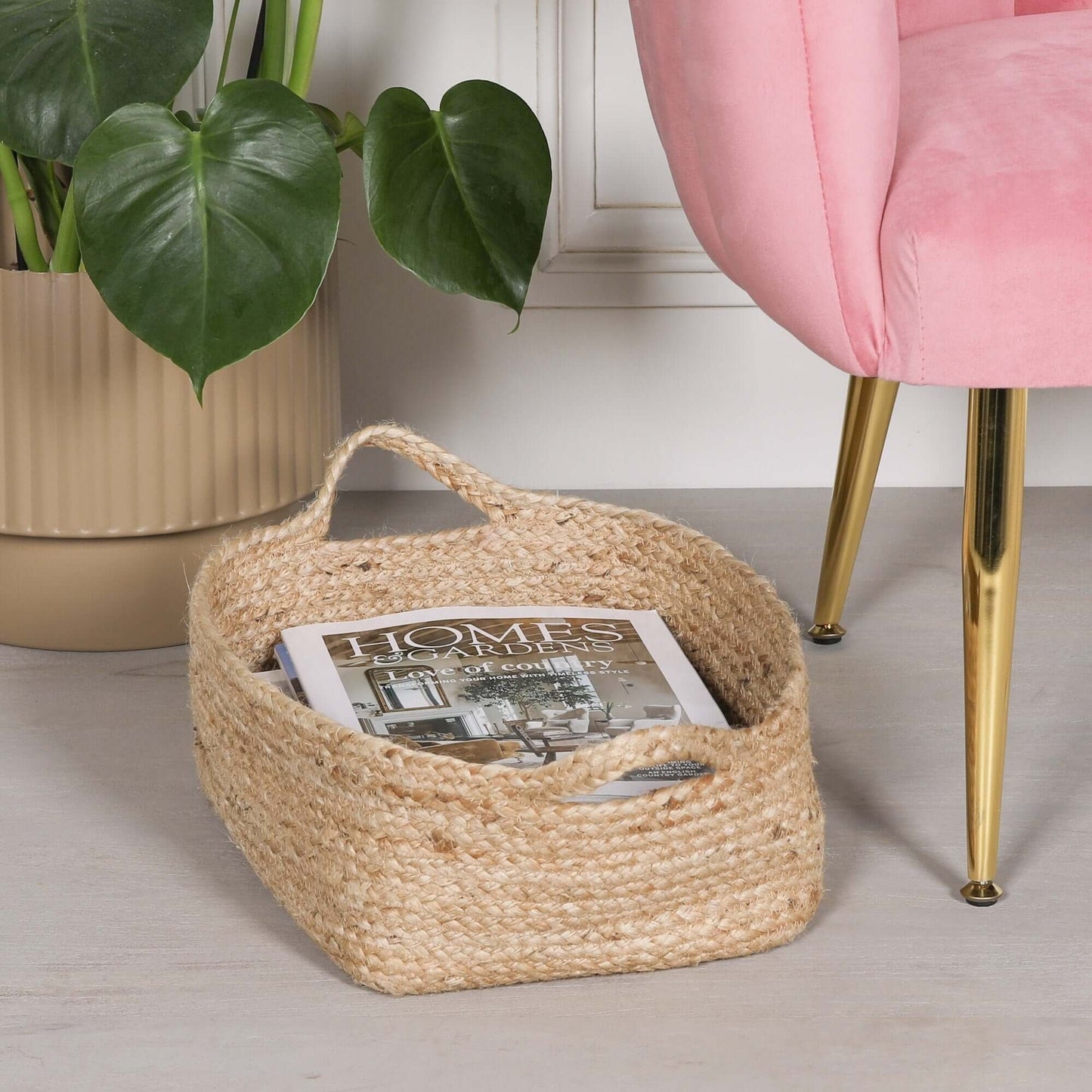 Jute Magazine Storage Basket holding a magazine, placed beside a pink chair and a potted plant.