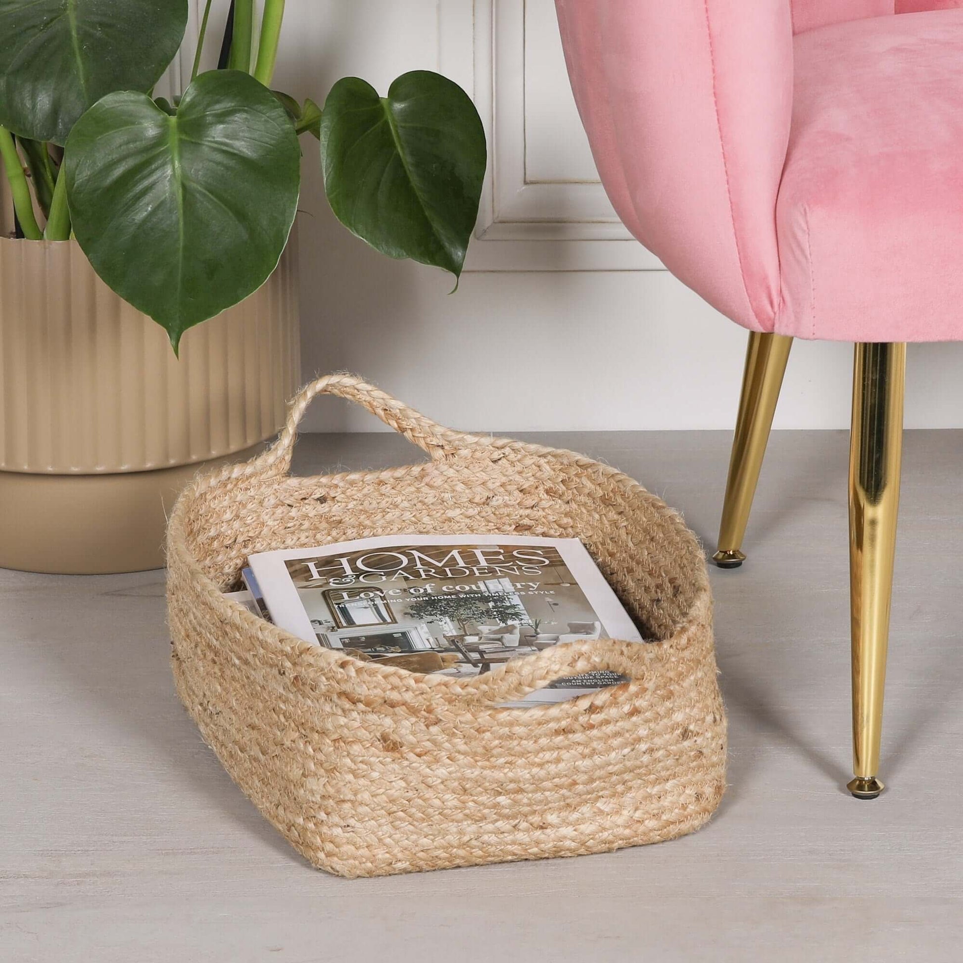 Jute Magazine Storage Basket holding a magazine, placed beside a pink chair and a potted plant.