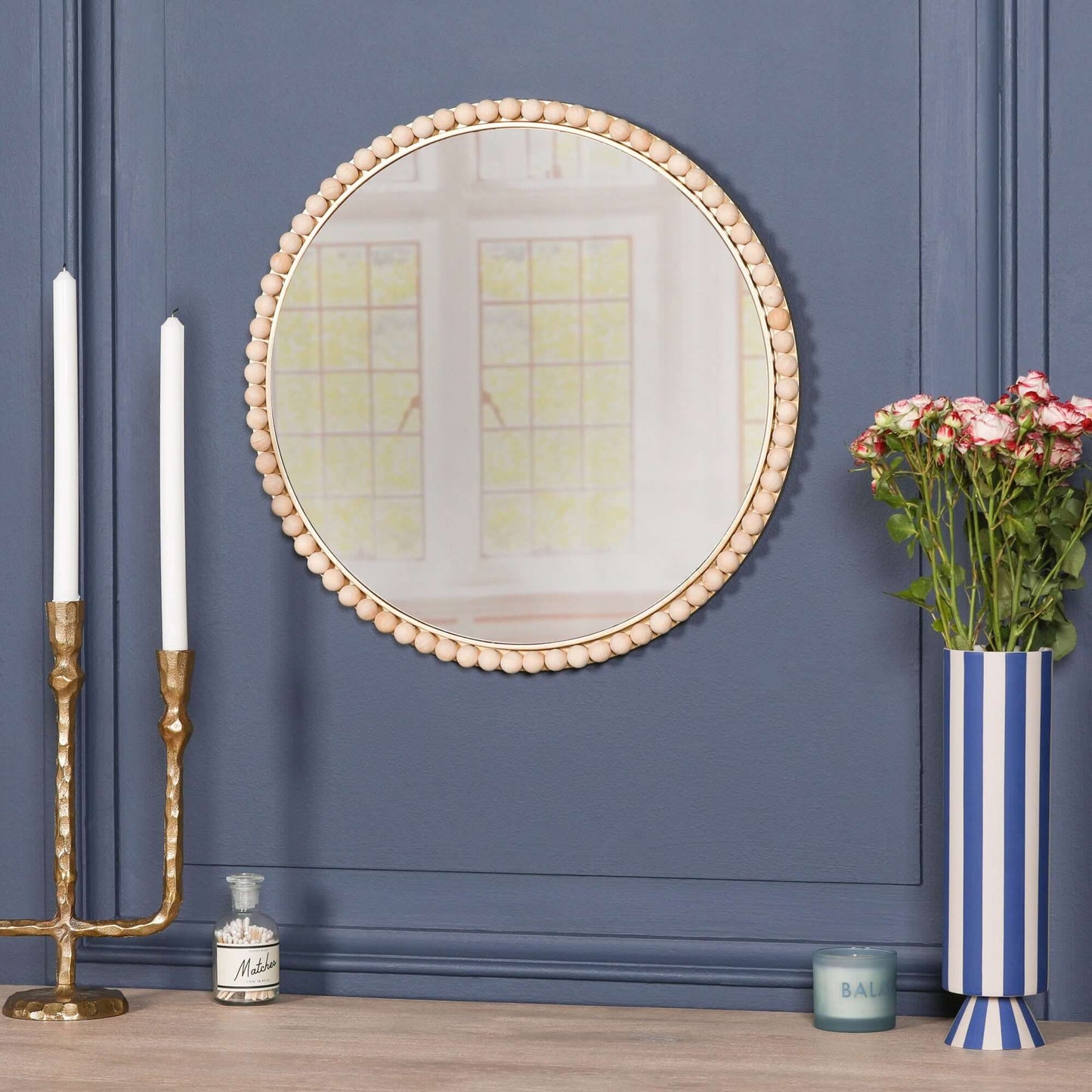 Wooden Bobble Round Wall Mirror displayed on a blue wall with candles and flowers in the background.